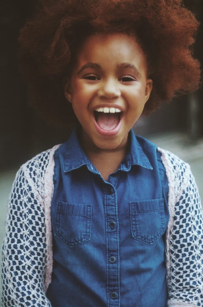 A cheerful young girl with afro hair wearing a denim shirt smiling outdoors.