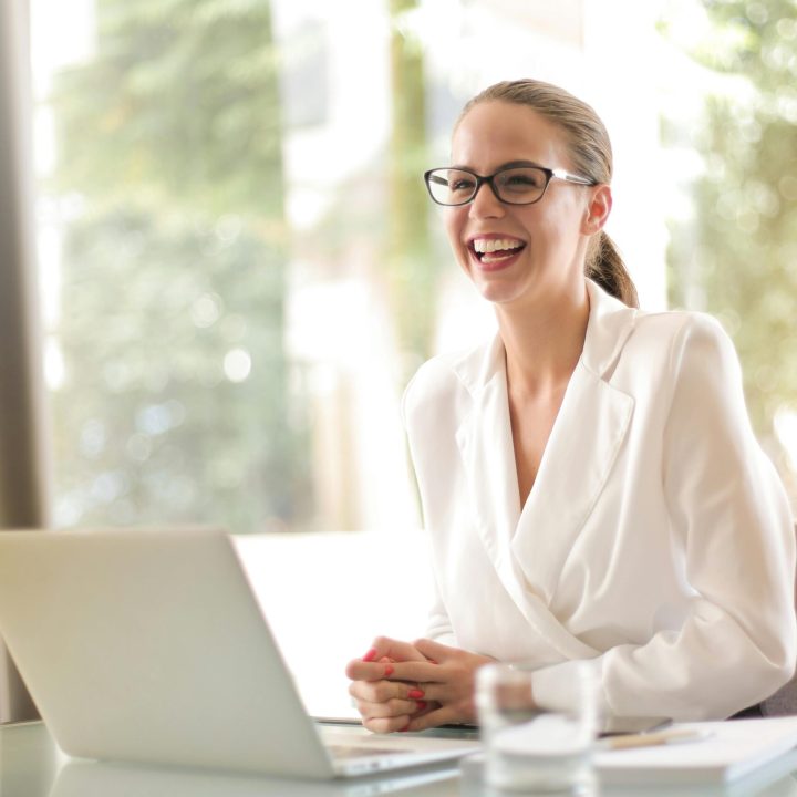 Cheerful businesswoman in glasses working on a laptop, in a bright and modern office setting.